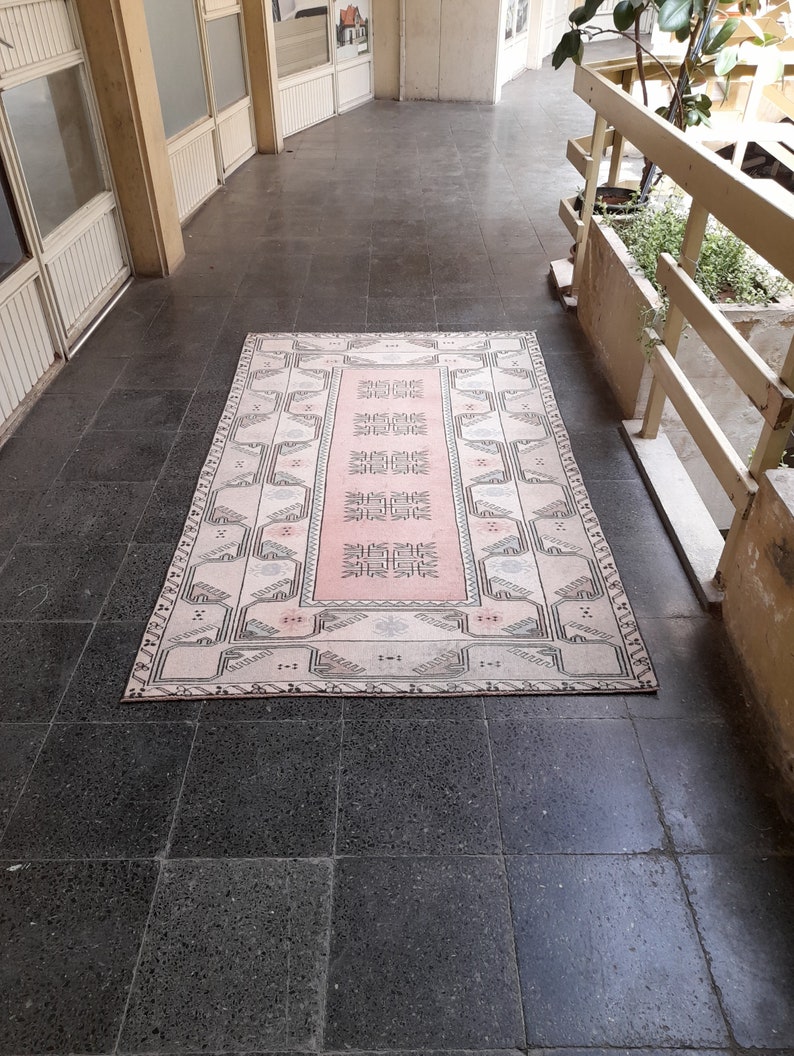 May include: A rectangular rug with a central pink panel and a cream border. The rug has a geometric pattern in pink, cream, and grey. It's placed on a dark grey tiled floor, with a building's exterior visible in the background. The rug's design is intricate.
