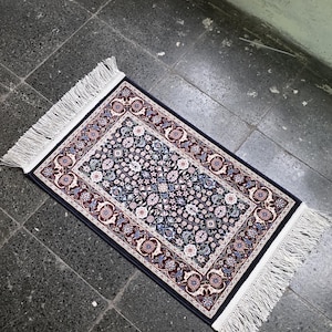 Op de afbeelding: Een rechthoekig tapijt met een donkerblauwe achtergrond en een bloemenpatroon in tinten wit, roze en blauw. Het tapijt heeft een decoratieve rand en witte franjes aan de korte uiteinden. Het tapijt is ongeveer 60 cm lang en 30 cm breed.