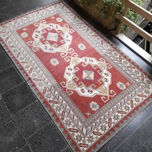 May include: A red, white, and beige patterned rug with a geometric design. The rug has a fringed edge and is made of wool.