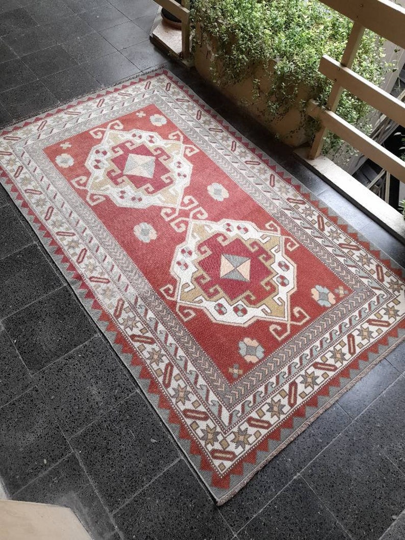 May include: A red, white, and beige patterned rug with a geometric design. The rug has a fringed edge and is made of wool.