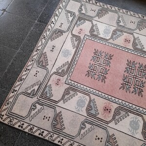 May include: A rectangular rug with a central pink panel and a cream-coloured border. The rug features geometric patterns in shades of grey, brown, and pink. The rug is placed on a dark grey tiled floor.