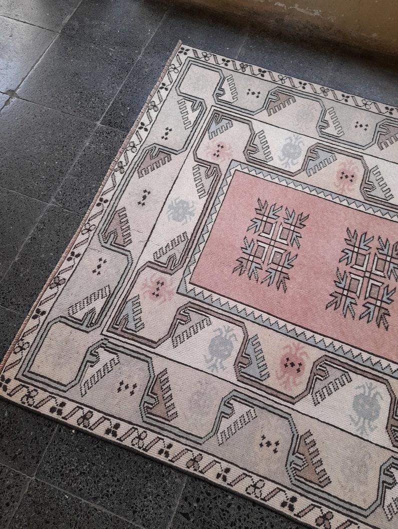 May include: A rectangular rug with a central pink panel and a cream-coloured border. The rug features geometric patterns in shades of grey, brown, and pink. The rug is placed on a dark grey tiled floor.