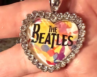 May include: A silver heart-shaped necklace with a clear glass pendant featuring a colourful floral design and the text "The Beatles". The pendant is surrounded by a border of clear rhinestones.