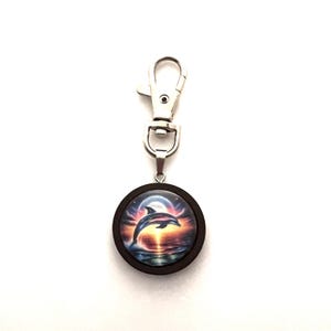 May include: A silver key chain with a round wooden charm featuring a colorful image of a dolphin jumping out of the water with a full moon in the background.