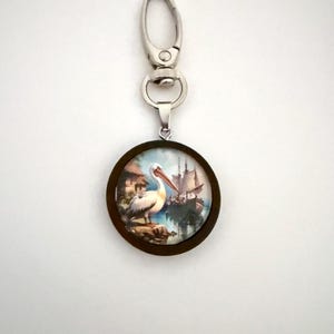 May include: A round keychain pendant with a nautical scene featuring a pelican and a ship. The pendant has a dark brown wooden frame and a silver-colored clasp.