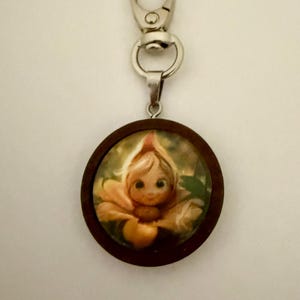 May include: A round wooden keychain featuring a whimsical illustration of a flower fairy with a smiling face. The fairy is depicted in shades of yellow and orange, set against a green background. The keychain has a silver clasp.