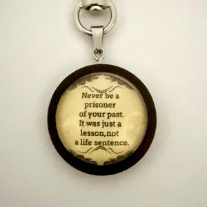 May include: A round keychain with a dark brown wooden frame and a silver-colored clasp. The center features text that reads, "Never be a prisoner of your past. It was just a lesson, not a life sentence."