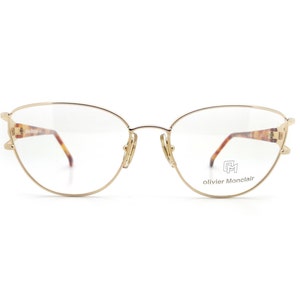 May include: Gold-tone metal eyeglasses with a cat-eye frame and clear lenses. The brand name "olivier Monclair" is printed on the right temple.