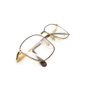 May include: Gold-tone metal eyeglasses with clear lenses. The glasses have a square frame with rounded edges.