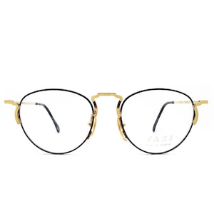 以下が含まれることがあります： 細い金色の金属フレームが付いた黒い眼鏡。右レンズにブランド名「TAXI」が印刷されています。