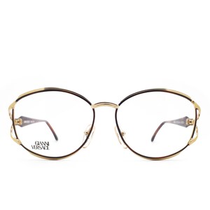 May include: Gold-tone oval-shaped eyeglasses with brown frames and clear lenses. The brand name "Gianni Versace" is printed on the left lens.