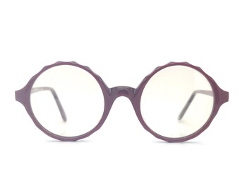 May include: A pair of round, burgundy-colored glasses with clear lenses. The frames have a scalloped edge.