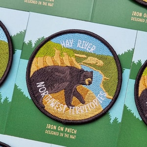 Peut inclure: Un écusson rond thermocollant représentant un ours noir marchant sur un paysage jaune et vert. Le texte "HAY RIVER NORTHWEST TERRITORIES" est brodé en blanc sur l'écusson. L'écusson est conçu dans les Territoires du Nord-Ouest.