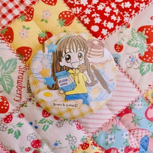 May include: A round, colorful button featuring a cartoon girl holding a box of Belgian waffles. The button also includes illustrations of eggs, a rolling pin, and a stack of pancakes. The text "Brunch time" is printed on the button.