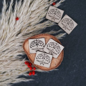 Peut inclure: Quatre étiquettes en bois avec un motif d'arbre noir et le texte "Tricote Main" sur une surface en bois brun. Les étiquettes sont entourées d'une plante blanche et moelleuse avec des baies rouges.