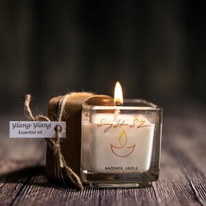 Peut inclure: Un bocal en verre carré contenant une bougie blanche. La bougie est étiquetée "Sunny Yellow SZ Handmade Candle Festival of Lights" et comporte un petit symbole de diya orange et jaune. La bougie est allumée et la flamme est visible. Un petit paquet brun avec une étiquette indiquant "Ylang-Ylang Essential Oil" se trouve au premier plan.