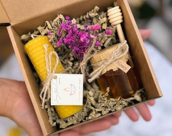 May include: A gift box containing a yellow beeswax candle, a small jar of honey with a wooden honey dipper, and a bundle of purple flowers. The candle has a textured surface and is tied with twine. The honey jar has a paper label that says "Honey". The box is filled with brown paper.