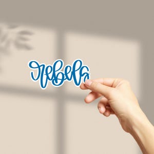May include: Blue vinyl sticker with the word "rebel" written in a cursive font.