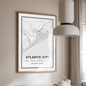 May include: A framed black and white map print of Atlantic City, New Jersey, with the coordinates 39°22'28"N / 74°23'44"W. The print is hanging on a light beige wall above a white table with a glass top. The table is surrounded by four wicker chairs.
