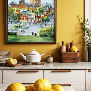 May include: A framed watercolor painting of a castle and cityscape hangs above a kitchen counter. A white teapot, lemons, and a wooden bowl with lemons are on the counter. The painting features a brown frame and a view of a city with a castle.