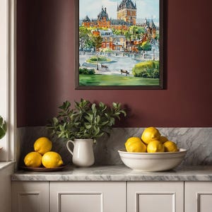 May include: A framed watercolor painting of a cityscape with a large castle-like building. The painting is displayed on a burgundy wall above a countertop with lemons in a bowl and on a plate, and a white pitcher with greenery.