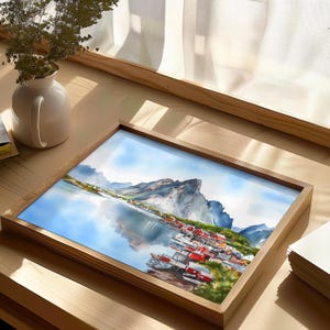 May include: A framed watercolor painting depicting a coastal village with red-roofed buildings nestled along a calm body of water, with a large mountain in the background. The artwork is set in a light brown wooden frame.