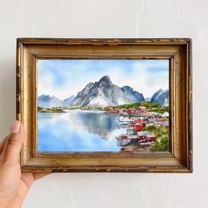 May include: Watercolor painting of a coastal village with red buildings, mountains, and a reflective lake. The artwork is framed in a gold, distressed wooden frame. The sky is blue with clouds.