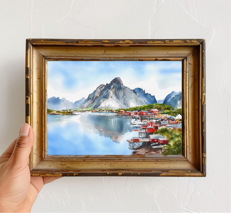 May include: Watercolor painting of a coastal village with red buildings, mountains, and a reflective lake. The artwork is framed in a gold, distressed wooden frame. The sky is blue with clouds.