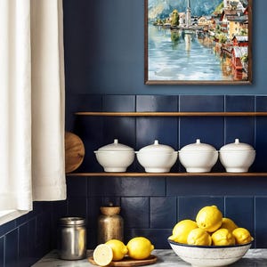 Peut inclure: Int&eacute;rieur de cuisine avec une aquarelle encadr&eacute;e d'un village au bord d'un lac. Des bols en c&eacute;ramique blanche avec couvercles sont sur une &eacute;tag&egrave;re au-dessus d'un comptoir en marbre. Un bol de citrons et une planche en bois sont sur le comptoir.