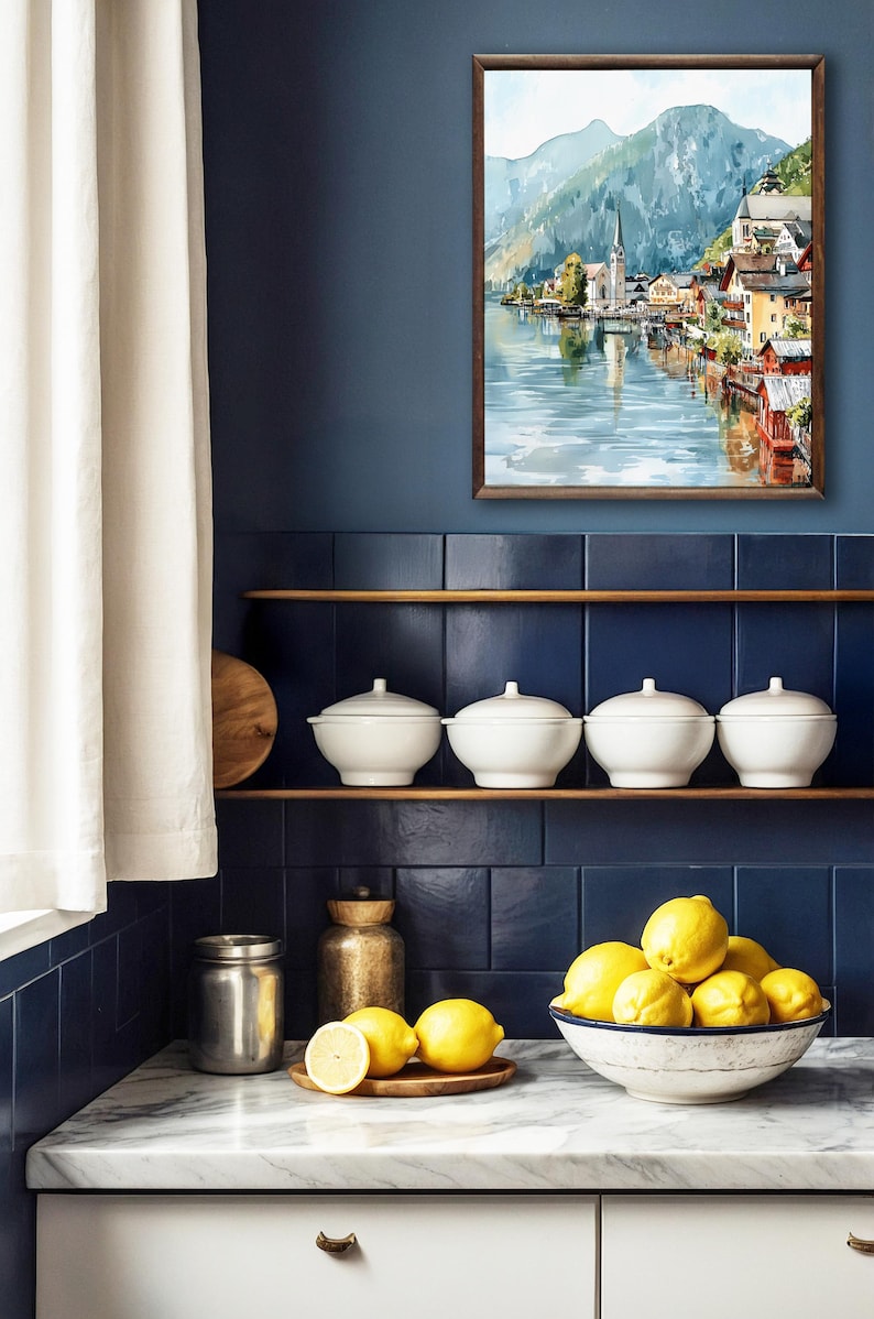 Peut inclure: Int&eacute;rieur de cuisine avec une aquarelle encadr&eacute;e d'un village au bord d'un lac. Des bols en c&eacute;ramique blanche avec couvercles sont sur une &eacute;tag&egrave;re au-dessus d'un comptoir en marbre. Un bol de citrons et une planche en bois sont sur le comptoir.