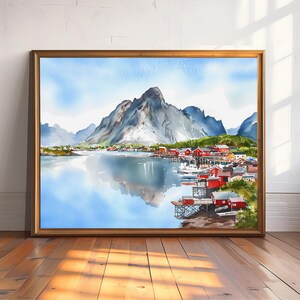 May include: Watercolor painting of a coastal village with red buildings, boats, and a mountain range reflected in the water. The artwork is framed in a gold-colored wooden frame and displayed on a wooden floor.