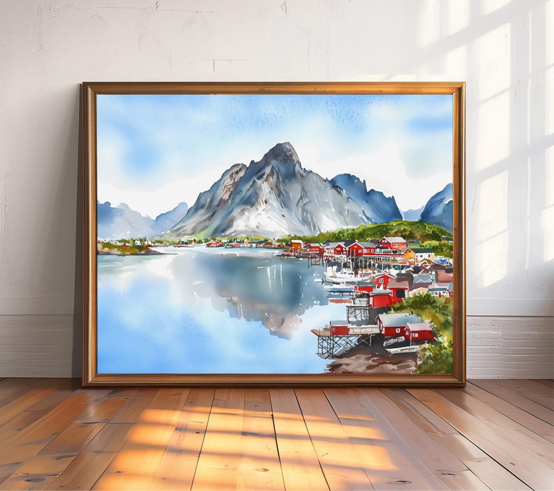 May include: Watercolor painting of a coastal village with red buildings, boats, and a mountain range reflected in the water. The artwork is framed in a gold-colored wooden frame and displayed on a wooden floor.