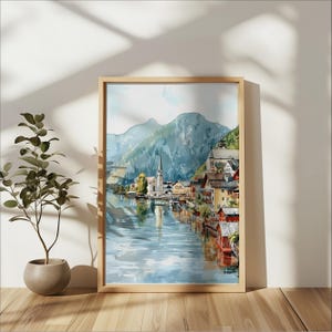 Peut inclure: Peinture &agrave; l'aquarelle d'un village europ&eacute;en au bord d'un lac, encadr&eacute;e dans un cadre en bois clair. L'&oelig;uvre repr&eacute;sente une &eacute;glise avec un haut clocher, des b&acirc;timents color&eacute;s et des montagnes. L'eau refl&egrave;te le village et le ciel.