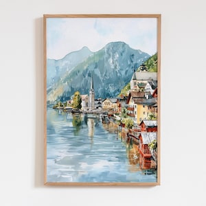 Peut inclure: Peinture &agrave; l'aquarelle d'un village au bord d'un lac avec un clocher d'&eacute;glise, des montagnes et des b&acirc;timents color&eacute;s. L'&oelig;uvre est encadr&eacute;e dans un cadre en bois clair, cr&eacute;ant une sc&egrave;ne sereine et pittoresque.