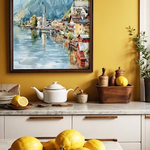 Peut inclure: Une peinture &agrave; l'aquarelle encadr&eacute;e d'un village au bord du lac avec une &eacute;glise et des montagnes. Une th&eacute;i&egrave;re blanche, des citrons et des bols en bois sont sur un comptoir de cuisine. Les citrons sont dans un bol en bois.