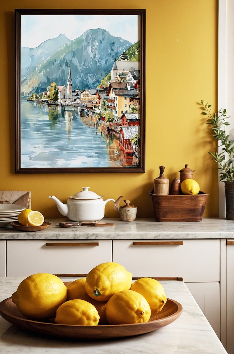 Peut inclure: Une peinture &agrave; l'aquarelle encadr&eacute;e d'un village au bord du lac avec une &eacute;glise et des montagnes. Une th&eacute;i&egrave;re blanche, des citrons et des bols en bois sont sur un comptoir de cuisine. Les citrons sont dans un bol en bois.