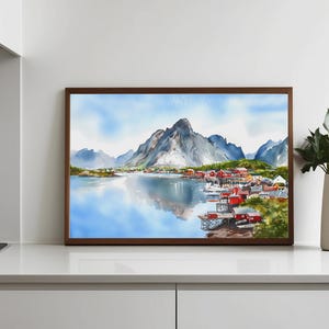 May include: Watercolor painting of a coastal village with red buildings, boats, and a mountain range reflected in the water. The artwork is framed in a dark brown wooden frame. The sky is a light blue with soft clouds.