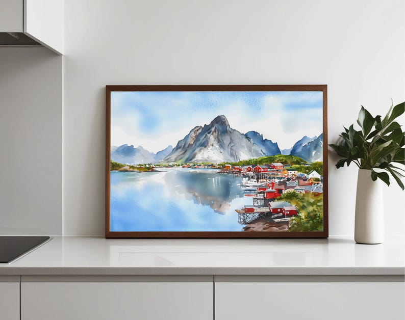 May include: Watercolor painting of a coastal village with red buildings, boats, and a mountain range reflected in the water. The artwork is framed in a dark brown wooden frame. The sky is a light blue with soft clouds.