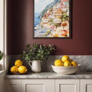 May include: A framed watercolor painting depicts a colorful coastal town nestled on a hillside. Below, a white pitcher with greenery and a bowl of lemons sit on a marble countertop, creating a vibrant kitchen scene.