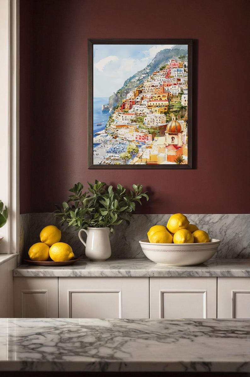 May include: A framed watercolor painting depicts a colorful coastal town nestled on a hillside. Below, a white pitcher with greenery and a bowl of lemons sit on a marble countertop, creating a vibrant kitchen scene.