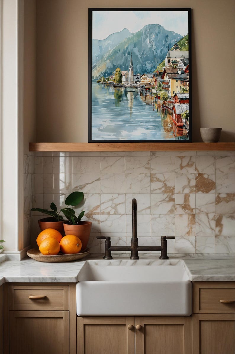Peut inclure: Une peinture &agrave; l'aquarelle encadr&eacute;e d'un village au bord du lac avec une &eacute;glise et des montagnes. Le tableau est accroch&eacute; au-dessus d'une &eacute;tag&egrave;re en bois. En dessous, un &eacute;vier de ferme blanc, des oranges et des plantes en pot.