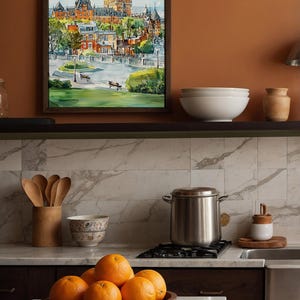 May include: A framed watercolor painting of a European city with a large castle. The painting is displayed above a kitchen shelf with white bowls and a ceramic jar. A wooden bowl of oranges sits on the counter.