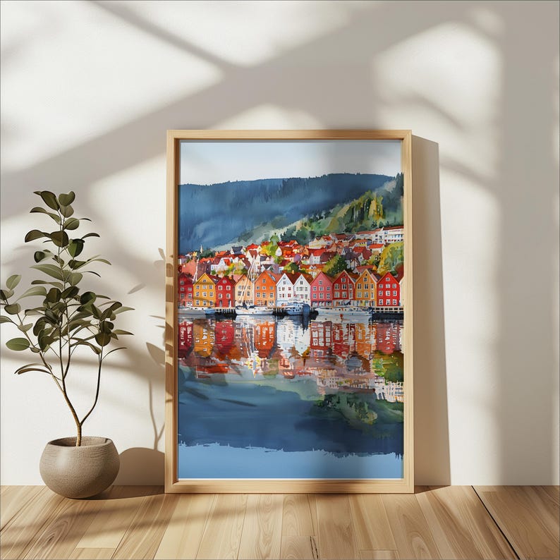 May include: Watercolor painting of a colorful waterfront town with buildings in shades of red, orange, yellow, and white. The artwork is framed in a light wood frame and reflects in the water below. A mountain range is in the background.