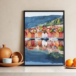 May include: Watercolor painting of a waterfront town with colorful buildings and boats. The buildings are reflected in the water, creating a symmetrical image. The artwork is framed in a black frame and is displayed on a neutral wall.