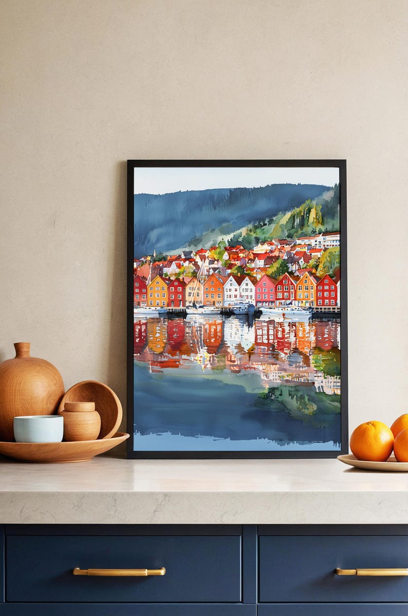 May include: Watercolor painting of a waterfront town with colorful buildings and boats. The buildings are reflected in the water, creating a symmetrical image. The artwork is framed in a black frame and is displayed on a neutral wall.