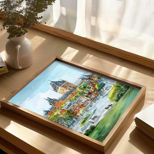May include: Watercolor painting of a European cityscape in a wooden frame. The artwork depicts buildings with orange, yellow, and brown roofs, surrounded by green trees and a blue sky. The frame is made of light-colored wood.
