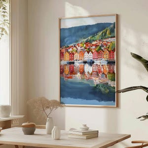 May include: A framed watercolor painting depicts a colorful waterfront scene. The artwork showcases a row of vibrant buildings in shades of red, orange, yellow, and white, reflected in the calm water below. A mountain range forms the backdrop.