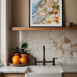 May include: A framed watercolor print of a coastal Italian town with colorful buildings and a domed church. The print is displayed above a wooden shelf. Below, a white farmhouse sink, oranges, and potted plants are arranged in a kitchen setting.
