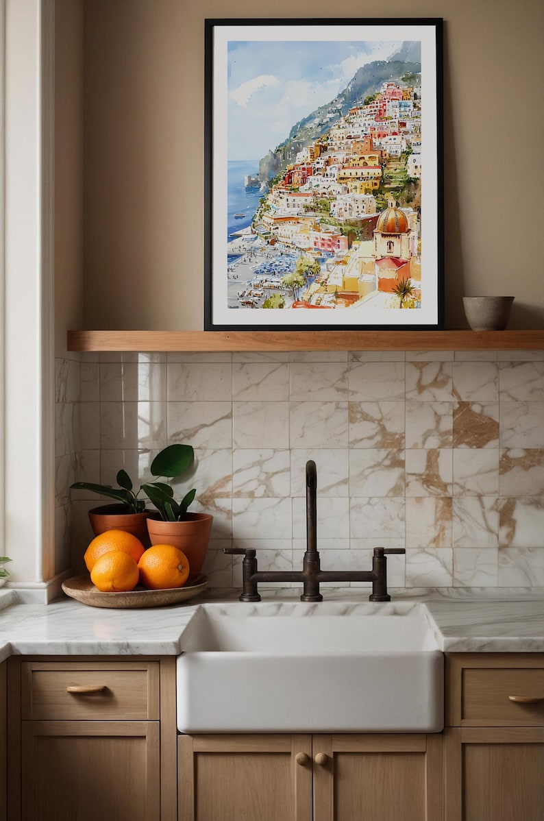 May include: A framed watercolor print of a coastal Italian town with colorful buildings and a domed church. The print is displayed above a wooden shelf. Below, a white farmhouse sink, oranges, and potted plants are arranged in a kitchen setting.