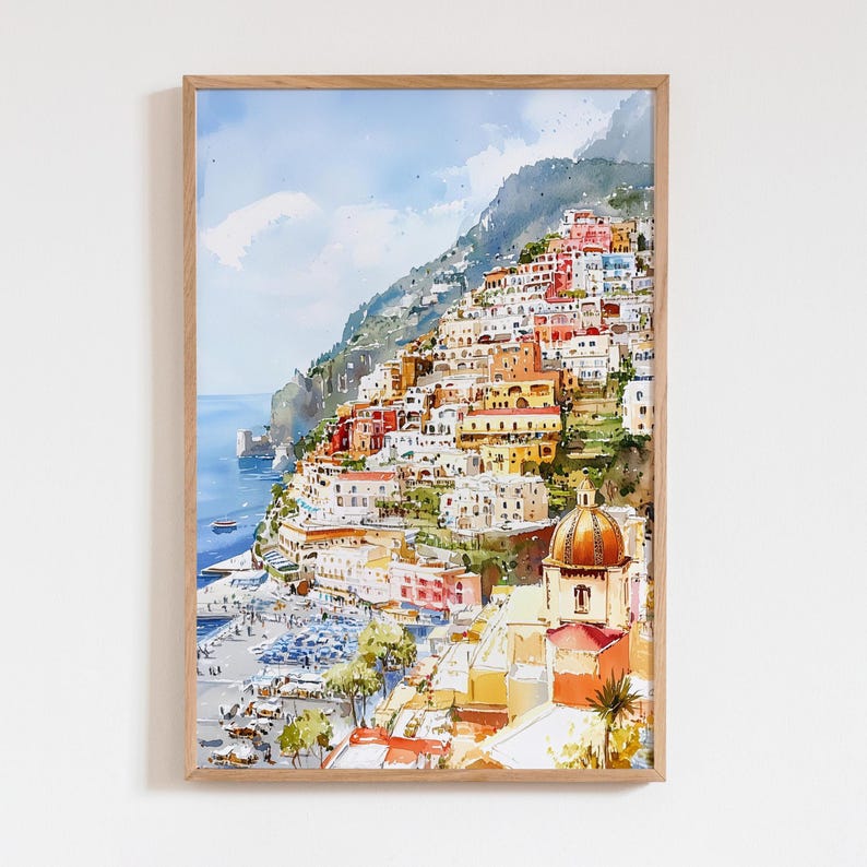 May include: Watercolor print of a coastal Italian town. The artwork depicts colorful buildings cascading down a hillside towards the sea. A church with a domed roof is visible. The print is framed in a light brown wooden frame.